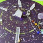 Crystal pendulum, calendar, and gemstones on a purple divination mat.  Various crystals, including points, tumbled stones, and a pyramid shaped pendulum.