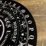 Black circular design, white lettering and symbols.  Mystical, occult style.  Possibly a magic or tarot themed coaster or mat.