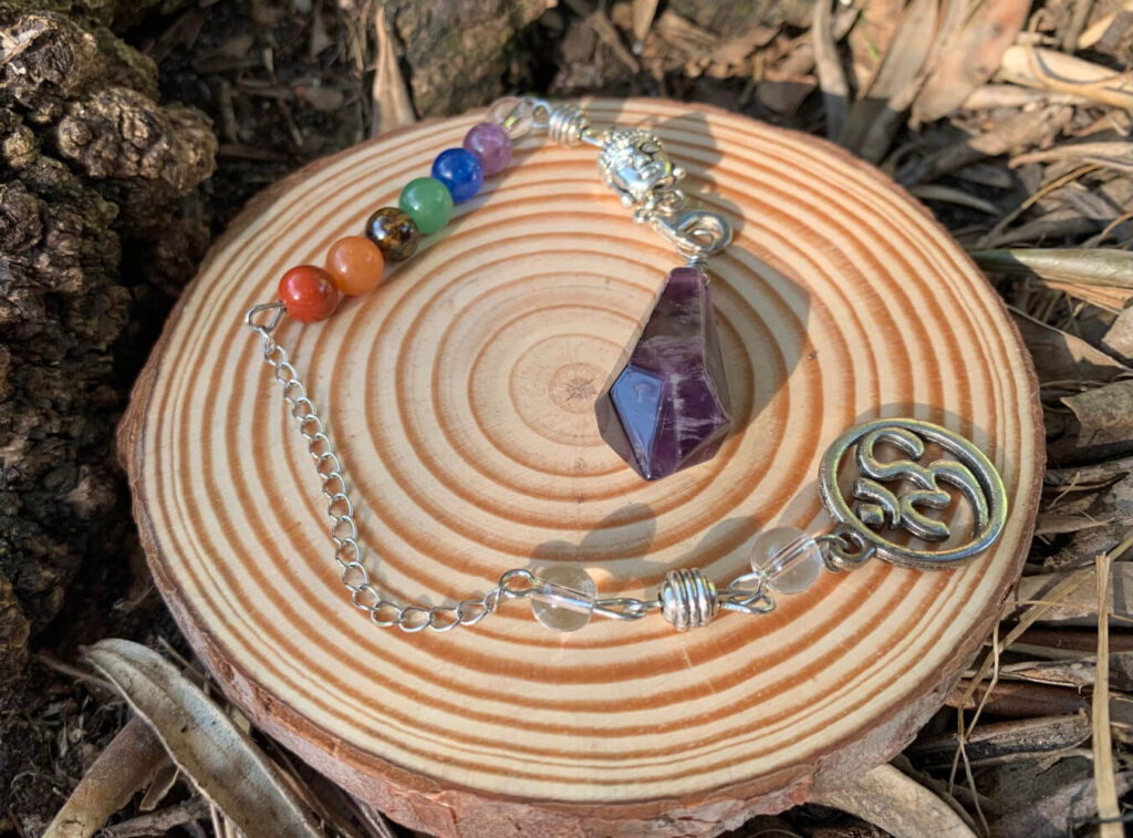 Rainbow chakra stones, amethyst pendulum, and Om symbol on wooden coaster.  Crystal therapy.