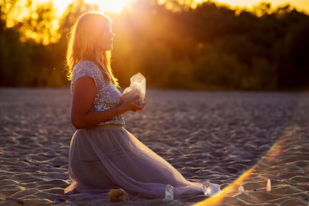 Woman kneeling on beach, holding crystals, sunset.