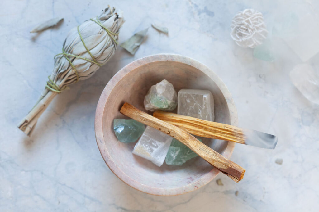 White sage smudge stick, crystals, and palo santo incense sticks in a stone bowl.
