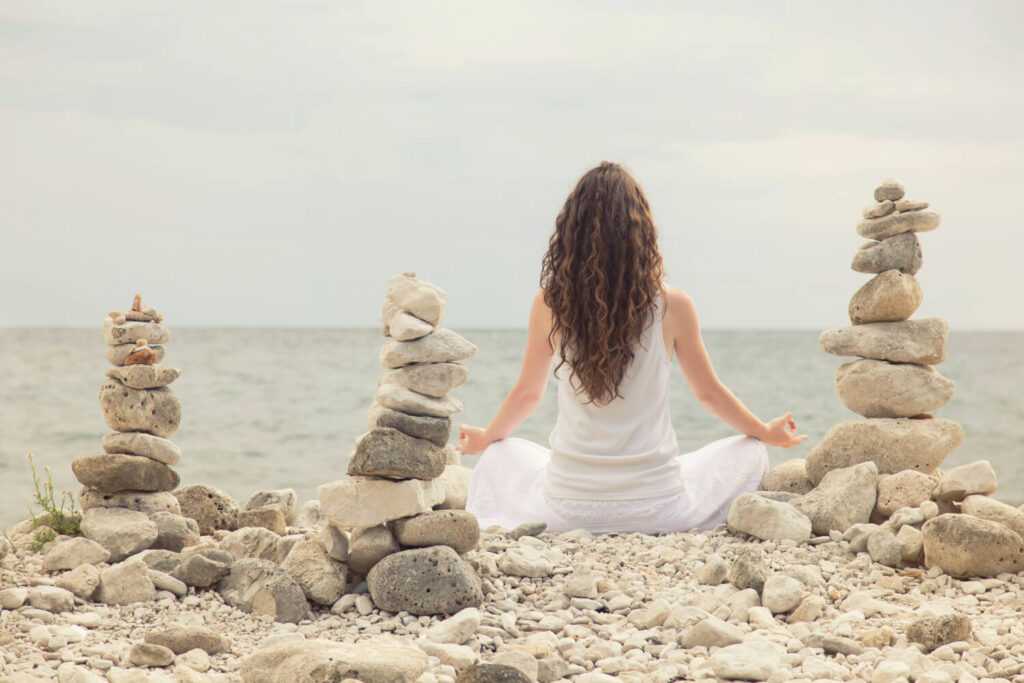 Woman in meditation pose on beach, surrounded by stacked stones.  Ocean view.