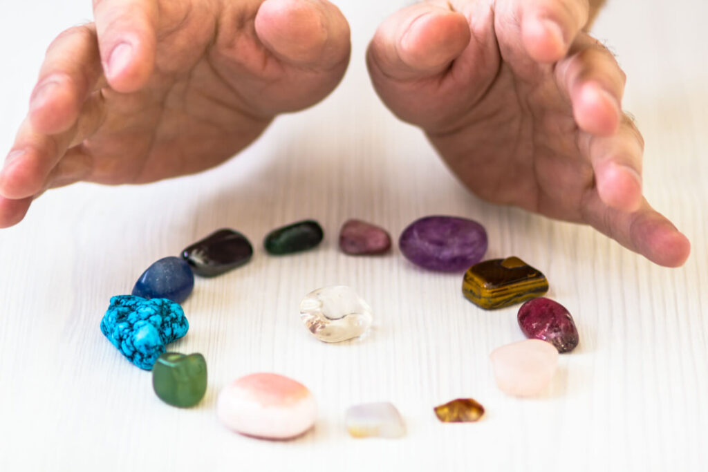 Hands holding various colored gemstones arranged in a circle on a white surface.