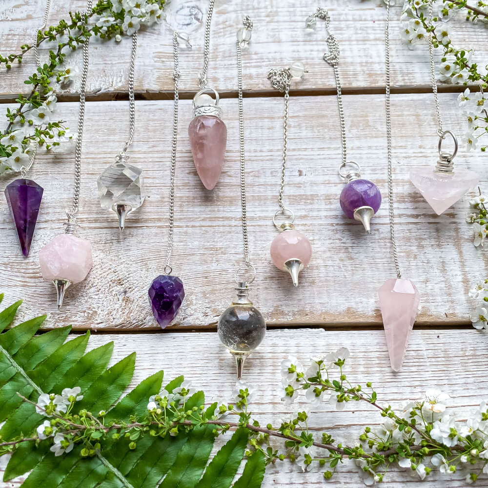 Various crystal pendulums, amethyst, rose quartz, clear quartz,  on a white wooden surface, with flowers and greenery.