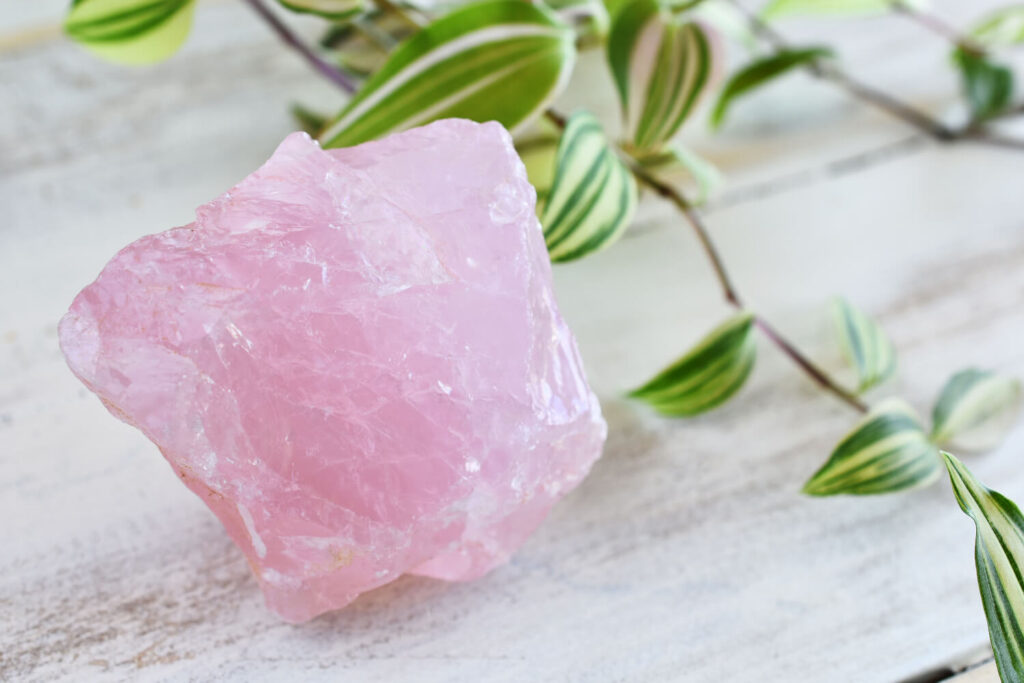 Pink raw rose quartz crystal on a white wooden surface with green leaves.