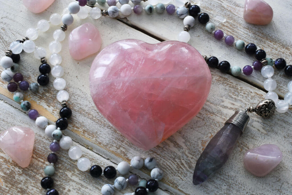Pink rose quartz heart,  crystal beads, amethyst pendant, and other stones on a light wooden surface.