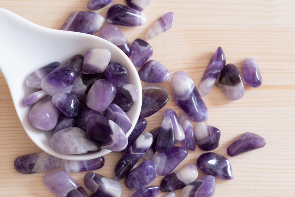 Small tumbled amethyst stones in a white bowl and scattered on light wood.