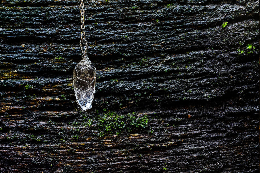 Clear quartz pendulum, wire-wrapped, hanging from chain, on dark, mossy wood.